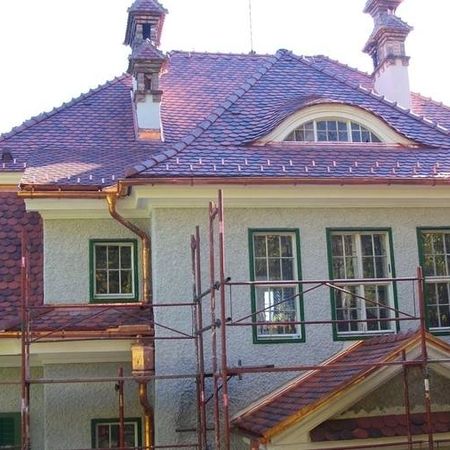 Vor einem Haus mit weißer Wand, grünen Fensterrahmen und rot / violettem Dach steht ein Baugerüst