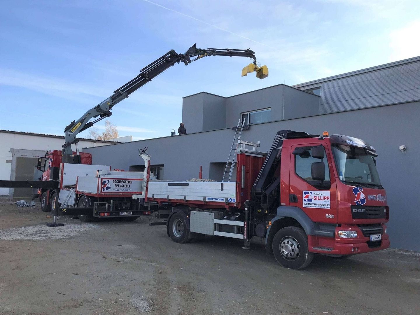 Zwei Fahrzeuge von der Friedrich Sillipp GmbH mit montierten Kran-Armen, einer ausgefahren und mit einem Greifer bestückt bearbeitet etwas auf einem Flachdach